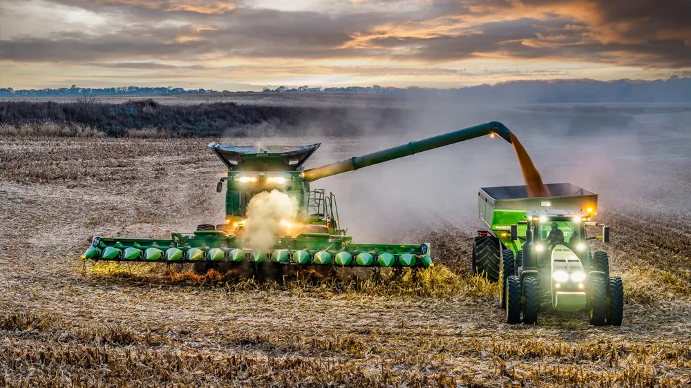 Aerial view of a combine unloading corn into a grain cart at dusk with dust and headlights lit